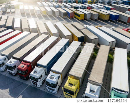 Aerial view of a large truck parking lot at a logistics hub, with multiple commercial semi trucks parked in organized rows Aerial view of a large truck parking lot at a logistics hub, with multiple commercial semi trucks parked in organized rows 127164234