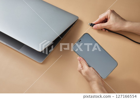 female hand connecting charger cable to laptop and holding smartphone at beige table, closeup female hand connecting charger cable to laptop and holding smartphone at beige table, closeup 127166554