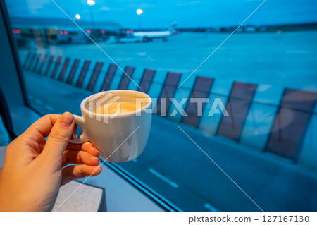 Hand holding coffee cup with night view of airport and planes in background. Concept of late-night flights, travel rituals, and quiet moments before departure 127167130