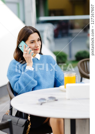 Young woman talking on smartphone and drinking orange juice at outdoor cafe 127167607