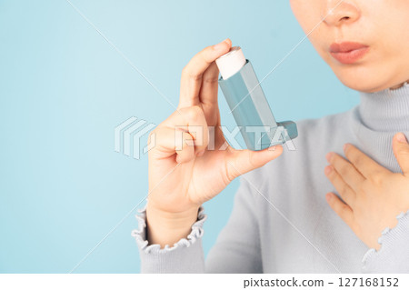 Young female using blue asthma inhaler for relief asthma attack. Pharmaceutical products is used to prevent and treat wheezing and shortness of breath caused asthma or COPD. Close up. Young female using blue asthma inhaler for relief asthma attack. Pharmaceutical products is used to prevent and treat wheezing and shortness of breath caused asthma or COPD. Close up. 127168152
