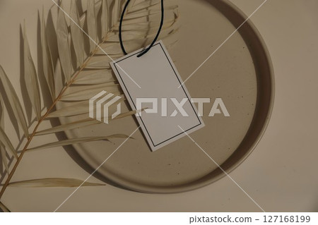 Minimalistic flat lay. Blank gift tag, label mockup on beige ceramic plate decorated with dried palm leaf. Neutral tones, soft shadows. Natural summer sale aesthetic design for branding. Flat lay, top 127168199