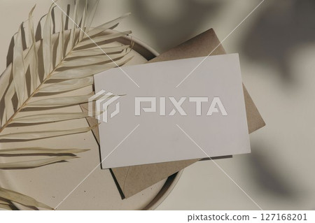 Blank horizontal greeting card, invitation mockup, craft envelope in sunlight, beige table background. Floral shadows overlay. Palm leaf on plate. Neutral web banner. Elegant stationery. Flat lay. Top 127168201