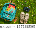 Open turquoise backpack with colorful school supplies, vintage camera, and sneakers on green grass, symbolizing back to school and outdoor learning 127168595