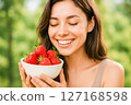 Happy young woman smiling with closed eyes, holding a bowl of fresh, ripe red strawberries, symbolizing healthy eating and natural enjoyment 127168598