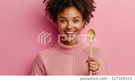 Cheerful young woman holding a wooden spoon against a pink background 127169523