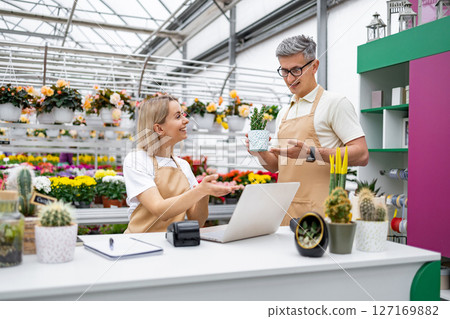 Two florists discuss a plant at the sales counter in a plant shop filled with various flowers and plants for sale. 127169882