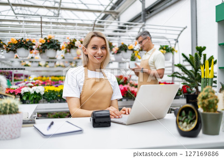 A florist is at work, smiling and using a laptop. In the background, a colleague is inspecting a plant, in a sunny greenhouse filled with flowers. 127169886