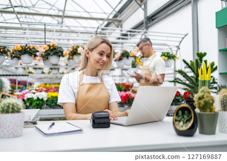 A smiling Caucasian florist uses a laptop behind a sale table, with another employee visible in the background. The shop is filled with flowers and plants. 127169887
