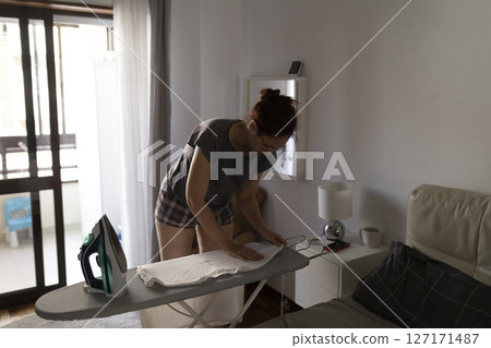 Woman ironing clothes on ironing board in bedroom 127171487
