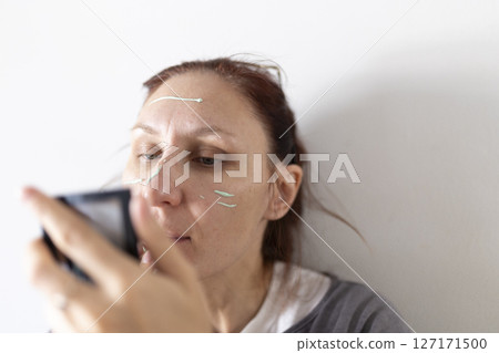 Woman applying green concealer cream on face with mirror 127171500