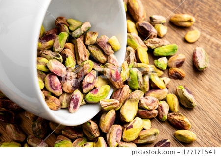 Shelled pistachio nuts spilled on wooden table from fallen white ceramic cup. Top view. Health food ingredient Shelled pistachio nuts spilled on wooden table from fallen white ceramic cup. Top view. Health food ingredient 127171541