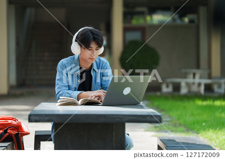 Portrait of a young Asian male college student e-learning using laptop on the bench on campus. 127172009