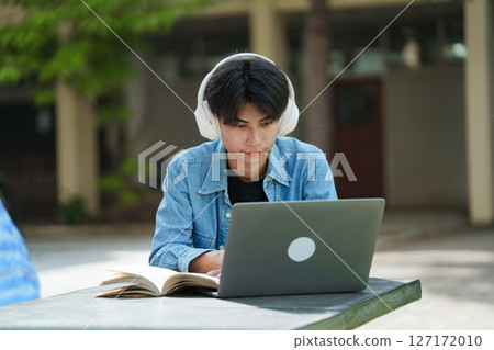 Portrait of a young Asian male college student e-learning using laptop on the bench on campus. 127172010
