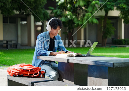 Portrait of a young Asian male college student e-learning using laptop on the bench on campus. Portrait of a young Asian male college student e-learning using laptop on the bench on campus. 127172013