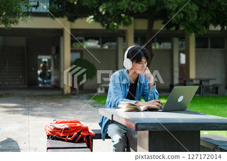 Portrait of a young Asian male college student e-learning using laptop on the bench on campus. 127172014