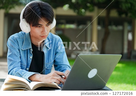 Portrait of a young Asian male college student e-learning using laptop on the bench on campus. 127172016