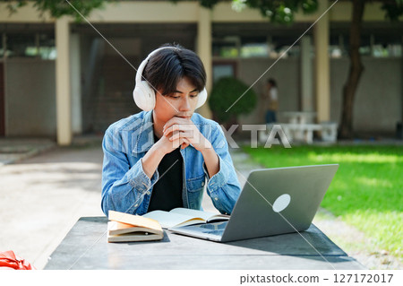 Portrait of a young Asian male college student e-learning using laptop on the bench on campus. 127172017
