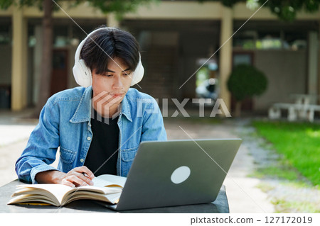 Portrait of a young Asian male college student e-learning using laptop on the bench on campus. 127172019