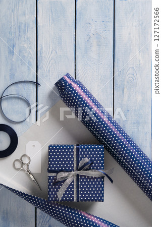 Gift wrapping items. Rolls of wrapping paper, scissors, ribbon, ready-wrapped gifts. Blue wooden background, space for your text. 127172566