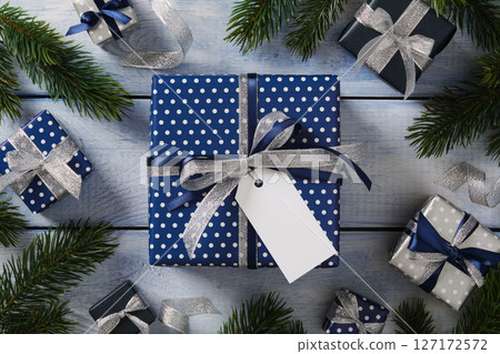 A blue gift with a silver ribbon and a blank tag for your message. Smaller gifts and fir branches surround it. 127172572