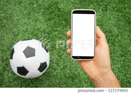 Man hand holds smartphone with white blank screen next to soccer ball on green grass. Online. Modern technology with traditional sports. Space for text. View from above. Empty display . 127174005