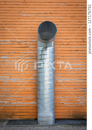 Tall Industrial Ventilation Pipe Against Weathered Orange Wooden Wall with Grate Base 127176792