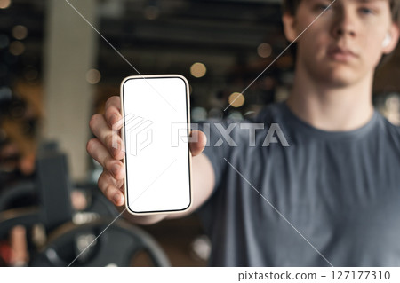 Athlete standing and displaying smartphone with white blank screen in gym setting. Mock up. Space for design. 127177310