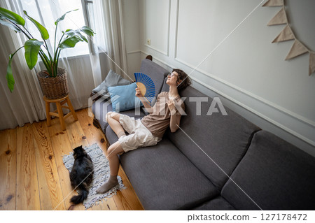 Sweaty woman using fan for relief, cat in heat exhaustion nearby. Home climate struggle, no AC 127178472