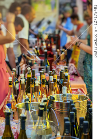Wine Festival. People gathered at busy wine tasting festival table filled with various bottles and ice buckets. Concept of celebration, public event, wine party Wine Festival. People gathered at busy wine tasting festival table filled with various bottles and ice buckets. Concept of celebration, public event, wine party 127178493