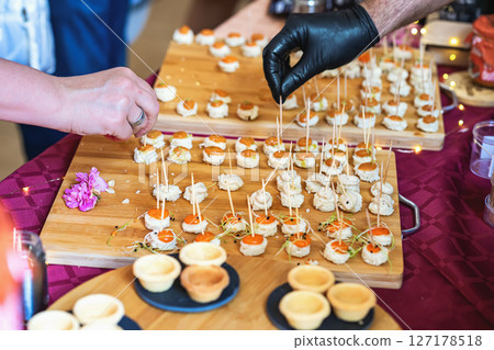 Hands placing gourmet appetizers with toothpicks on wooden trays at catering event. Concept of fine dining, finger food, festive culinary, catering, party food Hands placing gourmet appetizers with toothpicks on wooden trays at catering event. Concept of fine dining, finger food, festive culinary, catering, party food 127178518