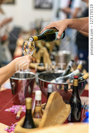 Wine tasting event with hand pouring white wine into glass, surrounded by wine bottles, ice buckets and wooden trays. Concept of celebration, sommelier service and premium lifestyle 127178519