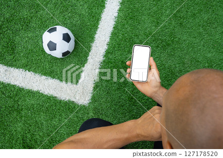 Man hand holds smartphone with white blank screen next to soccer ball on green grass. Online. Modern technology with traditional sports. Space for text. View from above. Empty display. Man hand holds smartphone with white blank screen next to soccer ball on green grass. Online. Modern technology with traditional sports. Space for text. View from above. Empty display. 127178520