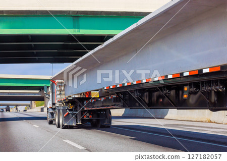 Truck carries long concrete slabs along highway surrounded by overpasses on clear day. Truck carries long concrete slabs along highway surrounded by overpasses on clear day. 127182057