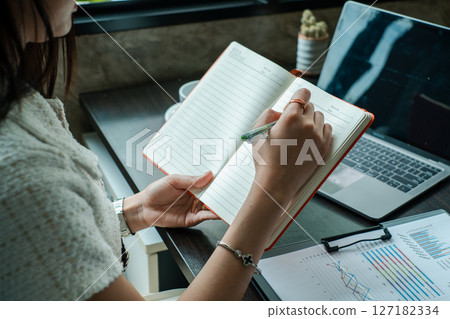 Professional woman taking notes at desk with laptop and financial documents, highlighting productivity and business planning. Professional woman taking notes at desk with laptop and financial documents, highlighting productivity and business planning. 127182334