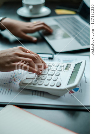 Close-up of hands using a calculator and laptop, analyzing financial data in a modern office environment. 127182336