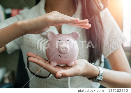 Close-up of a woman holding and protecting a pink piggy bank, representing savings and financial security in a modern environment. 127182350