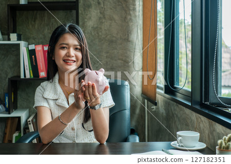 Smiling woman in office holding a piggy bank, symbolizing savings and finance, with books and coffee on the desk. Smiling woman in office holding a piggy bank, symbolizing savings and finance, with books and coffee on the desk. 127182351