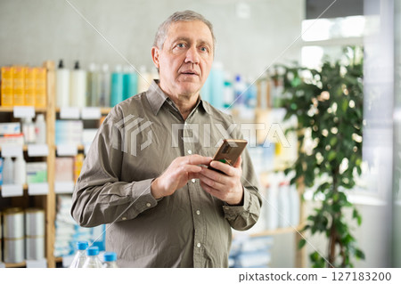 elderly man holds a mobile phone in his hands against the background of a counter in a pharmacy elderly man holds a mobile phone in his hands against the background of a counter in a pharmacy 127183200
