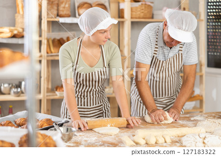 Young girl helps an elderly baker prepare raw dough - rolling it out with rolling pin Young girl helps an elderly baker prepare raw dough - rolling it out with rolling pin 127183322