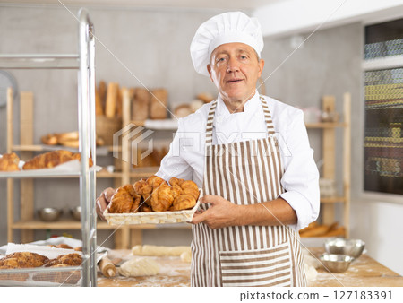 Bakery mature man employee holds basket of ready-made croissants. Bakery mature man employee holds basket of ready-made croissants. 127183391