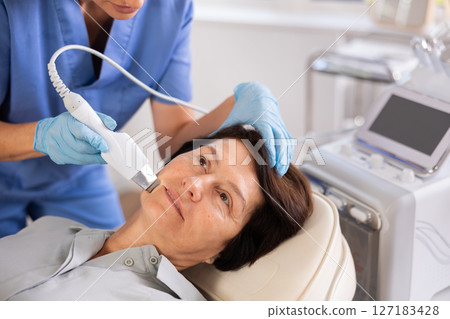 Mature woman undergoing hardware cosmetic facial treatment using ultrasonic shovel 127183428
