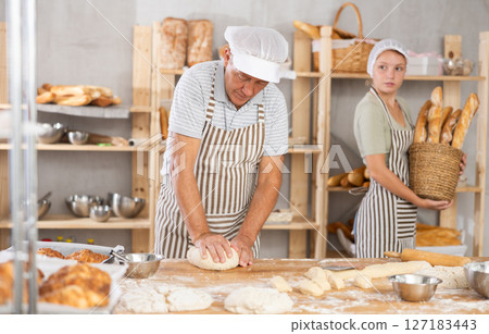 Elderly baker stands at his work bench, kneading and shaping dough to make bread, croissants and baguettes Elderly baker stands at his work bench, kneading and shaping dough to make bread, croissants and baguettes 127183443