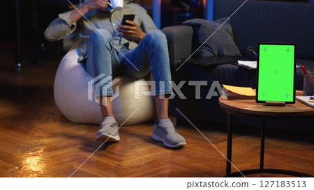 Black young person browses laptop and phone next to green screen, feeling casual and relaxed on the bean bag in his living room. Happy chill man in his trendy loft with gadgets. Camera B. Black young person browses laptop and phone next to green screen, feeling casual and relaxed on the bean bag in his living room. Happy chill man in his trendy loft with gadgets. Camera B. 127183513