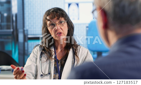 Senior woman evaluating diagnostic results with her medic in a cabinet, using advanced tech tools to give the correct results to his patient. Care management within medical industry. Camera B. Senior woman evaluating diagnostic results with her medic in a cabinet, using advanced tech tools to give the correct results to his patient. Care management within medical industry. Camera B. 127183517