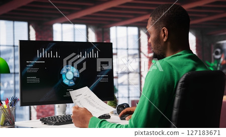 Black freelancer at home watching business conference on PC monitor, looking over statistics charts. Remote worker in apartment office follows presentation graphs on computer. Camera A. 127183671