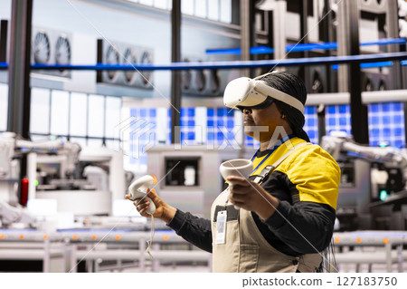 Factory worker wearing VR headset and holding motion controllers engages in virtual reality training in advanced industrial plant. Female engineer highlighting the role of AR in modern manufacturing. 127183750