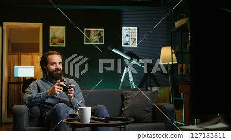 Joyful gamer at home gaming and enjoying leisure time. Smiling man sitting on couch in neon lit living room, excited to play videogame on console, relaxing after hard day at work, camera B 127183811
