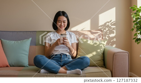 Happy Young Woman Using Smart phone While Relaxing on Cozy Sofa at Home 127187332