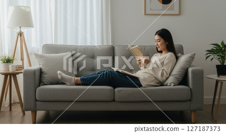 Young East Asian Woman in Cozy Lounge wear Reading on a Modern Grey Sofa in Minimalist Living Room 127187373